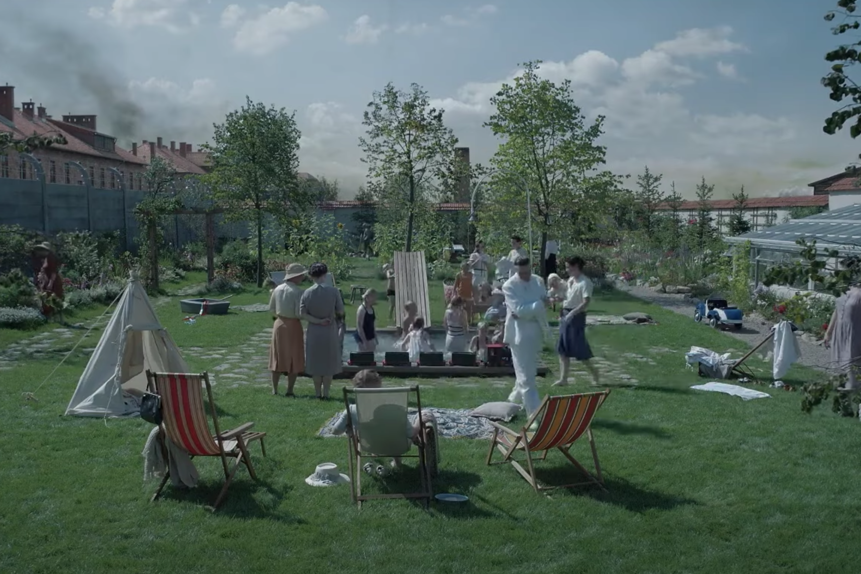 Una scena dal film 'La zona di interesse' mostra un giardino ben curato in un ambiente domestico. Persone, tra cui donne e bambini, si godono una giornata all'aperto. Alcuni bambini giocano nell'acqua di una piccola vasca, mentre adulti li osservano. In primo piano si vedono sedie a sdraio, una tenda e oggetti personali sparsi sul prato. Sullo sfondo, una fila di edifici con un muro di cemento e camini da cui fuoriesce fumo nero suggerisce una presenza inquietante, in contrasto con la serenità del giardino.