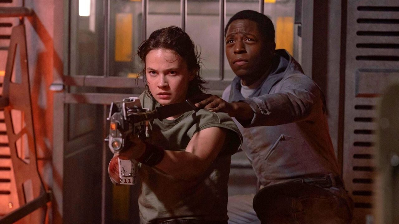 Questa immagine tratta dal film “Alien Romulus” ritrae due personaggi in un momento di tensione e di azione. Rain in camicia verde è in primo piano e impugna una grande arma futuristica, puntandola con grande attenzione. Dietro di lei, l'androide Andy una giacca chiara si accovaccia vicino a lei, con la mano sulla spalla, per guidarla o sostenerla. L'ambientazione sembra essere in un ambiente industriale o spaziale poco illuminato, con pareti metalliche e un senso di urgenza. Le espressioni e la postura dei personaggi suggeriscono che si stanno preparando per un confronto, forse con una pericolosa minaccia aliena.