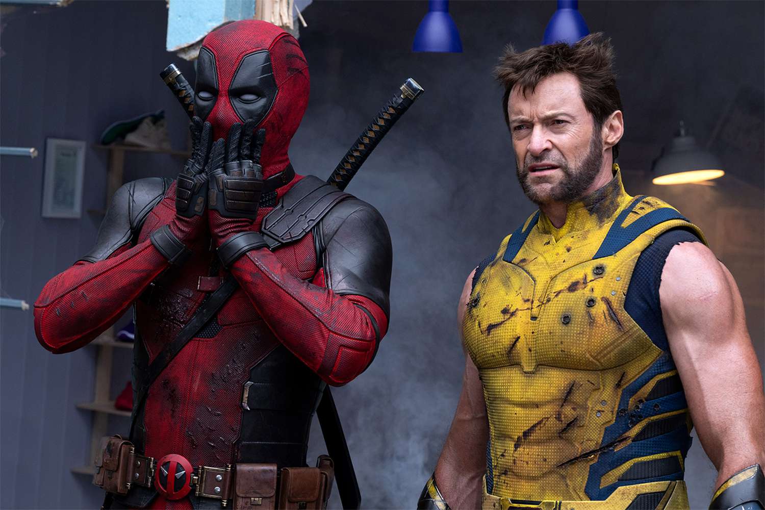 In una scena del film "Deadpool e Wolverine", Deadpool, vestito con la sua iconica tuta rossa e nera con due spade sulla schiena, sta in piedi con le mani che coprono il suo volto mascherato per l'apparente sorpresa. Accanto a lui, Wolverine indossa una robusta armatura gialla e blu, visibilmente consumata da graffi e sporco. Wolverine ha un'espressione intensa e concentrata sul volto, le braccia muscolose esposte e la sua caratteristica peluria facciale e acconciatura scompigliata. I due si trovano in mezzo ai detriti di quella che sembra essere una caotica scena di battaglia, evidenziata da una luce fioca e drammatica sullo sfondo.