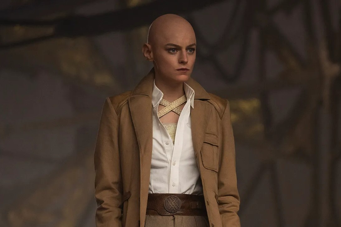 Questa immagine tratta dal film “Deadpool e Wolverine” ritrae Emma Corrin nei panni di Cassandra Nova, che indossa un trench marrone chiaro su una camicia bianca che presenta un particolare disegno di cinghie incrociate vicino al colletto. Il personaggio ha un'espressione severa e determinata, in piedi in un ambiente industriale poco illuminato. L'abbigliamento è accessoriato con un'ampia cintura marrone con un emblema circolare al centro, che contribuisce alla presenza imponente del personaggio. La scena suggerisce un momento di intensità o di confronto, con il personaggio che appare risoluto e pronto all'azione.