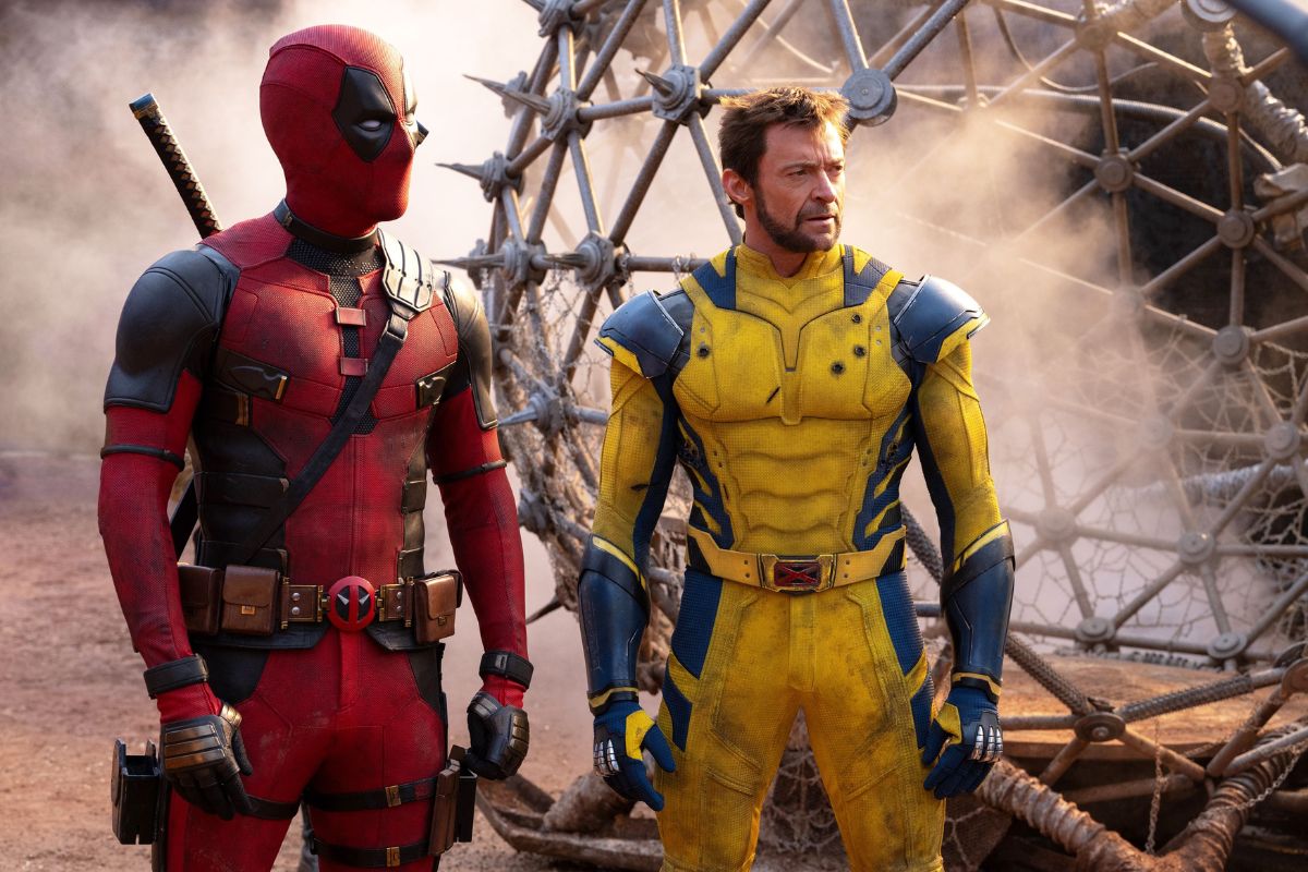 Questa immagine tratta dal film “Deadpool e Wolverine” mostra i due iconici personaggi fianco a fianco in un ambiente polveroso all'aperto. Deadpool, vestito con il suo caratteristico abito rosso e nero e con una katana legata alla schiena, è in piedi con le mani sui fianchi. Accanto a lui c'è Wolverine, che indossa una tuta gialla e blu che ricorda il suo classico costume dei fumetti. Gli artigli di Wolverine non sono estesi ed entrambi i personaggi hanno un'espressione seria mentre guardano in lontananza. Sullo sfondo si staglia una grande struttura metallica dai dettagli intricati, che aggiunge un elemento drammatico alla scena. L'immagine cattura un momento di intensità e attesa tra i due supereroi.