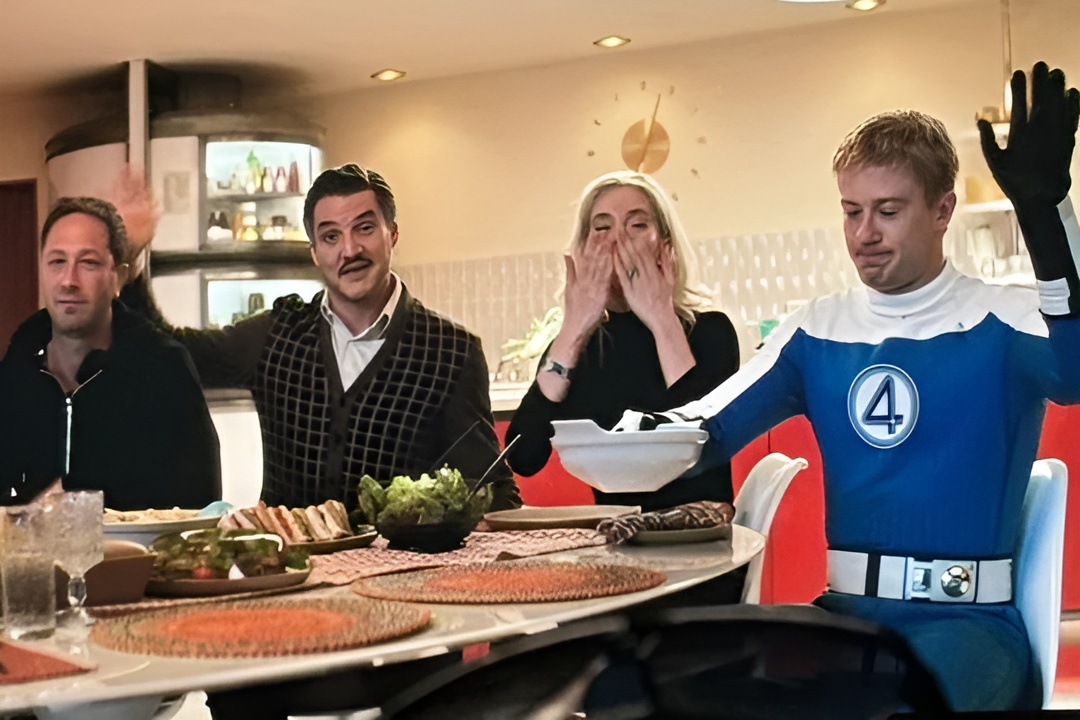 Un'immagine del dietro le quinte dal set di "Fantastic 4: First Steps". La scena mostra quattro persone sedute intorno a un tavolo da pranzo in una cucina dallo stile retrò. Una persona indossa un'uniforme blu dei “Fantastici 4” con l'iconico emblema del “4” e alza una mano guantata. Un altro individuo, vestito in modo casual, saluta allegramente, mentre una donna vestita di nero manda un bacio e un uomo con un gilet a fantasia sorride alla telecamera. La tavola è apparecchiata con vari piatti, tra cui panini, insalata e bicchieri di vetro. L'illuminazione calda e l'arredamento vintage evocano un'estetica anni '60, sottolineando il tono nostalgico del film.