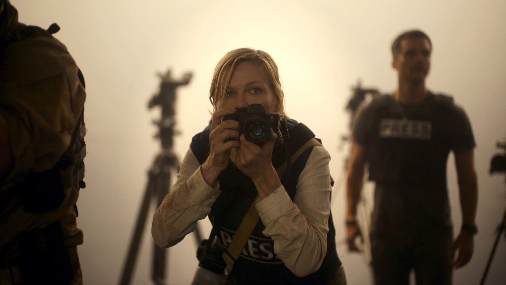 Questa immagine tratta dal film “Civil War” mostra una donna in primo piano, che indossa un giubbotto stampa e tiene in mano una macchina fotografica, mentre cattura un momento con un'attenzione intensa. Lo sfondo è nebuloso, con un gruppo di persone e attrezzature fotografiche leggermente visibili, che aggiungono un senso di caos o di urgenza. La scena fa pensare a una zona di conflitto o a un'area di disordini, con l'espressione determinata della donna che sottolinea la gravità della situazione che sta documentando. L'illuminazione è tenue, con una tonalità dorata che sottolinea l'atmosfera di tensione e azione.