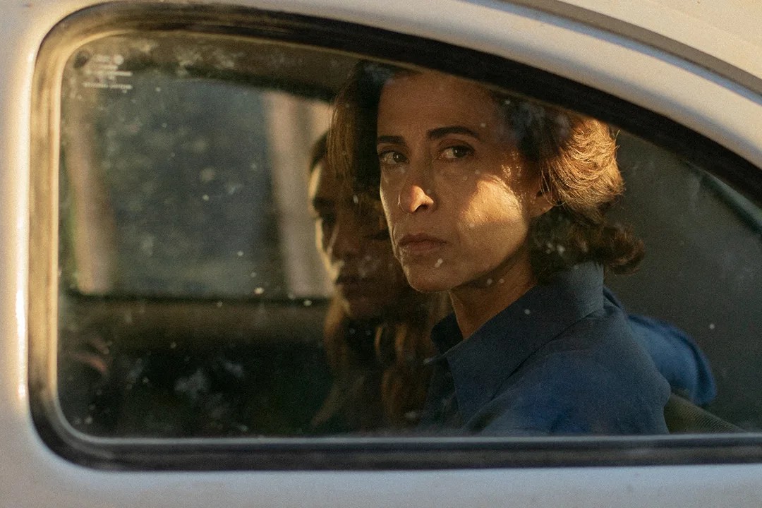 Una donna dall'espressione seria e contemplativa è vista attraverso il finestrino polveroso di un'automobile. Un'altra persona, parzialmente visibile, è seduta accanto a lei. La scena cattura un momento intenso e riflessivo, con l'illuminazione naturale che mette in risalto i personaggi, dal film 'Io sono ancora qui'.