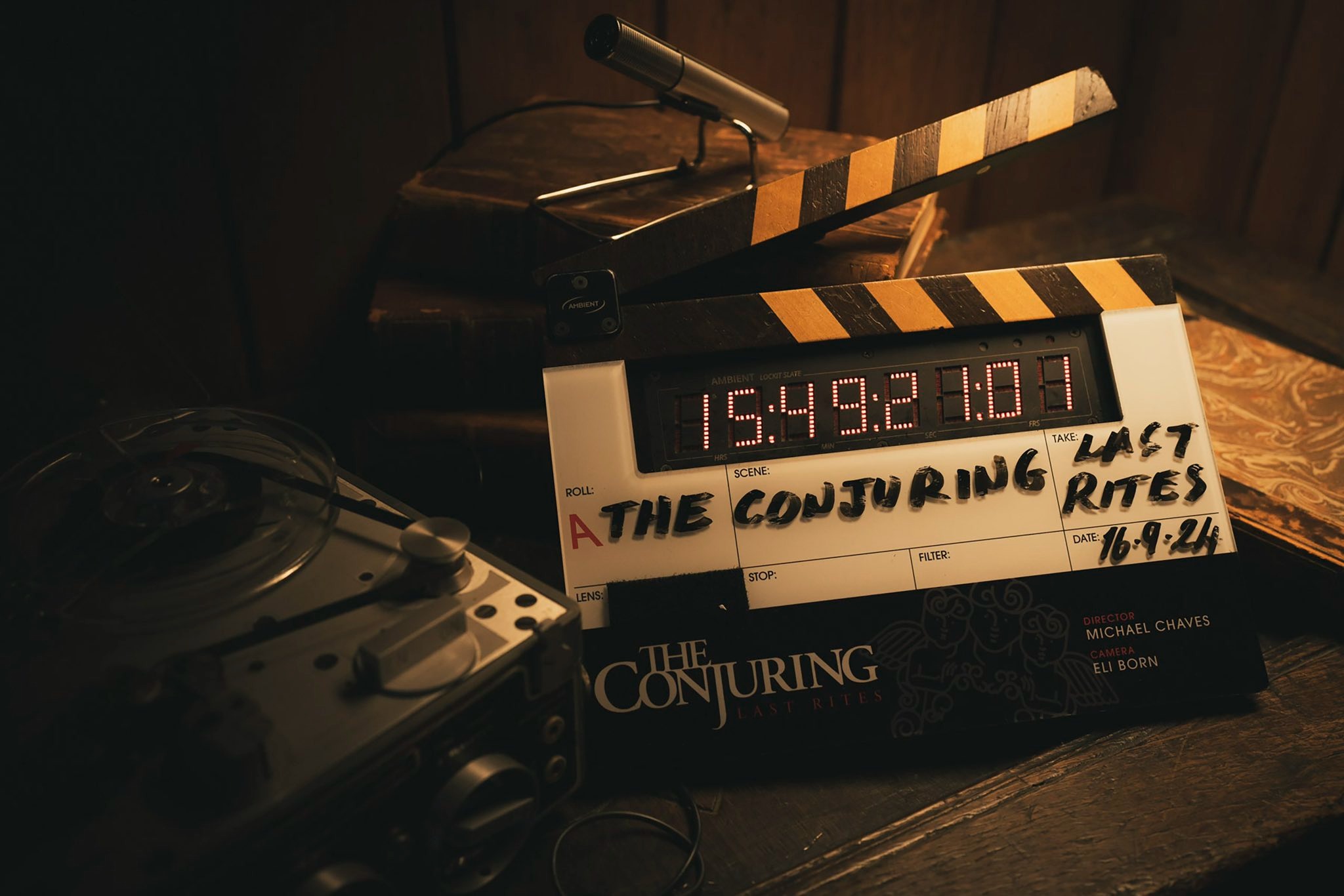 Un'immagine del dietro le quinte dal set di "The Conjuring 4: Last Rites". L'attenzione si concentra su un ciak etichettato con il titolo del film, “The Conjuring: Last Rites” e la data ‘16.9.24’. Il ciak è appoggiato su un tavolo di legno, circondato da apparecchiature di registrazione vintage, tra cui un registratore a bobine e un microfono classico. Un'illuminazione fioca e lunatica esalta l'atmosfera inquietante, con ombre e toni caldi che contribuiscono al tema soprannaturale del film. Sullo sfondo, vecchi libri e superfici strutturate contribuiscono all'estetica vintage e inquietante.