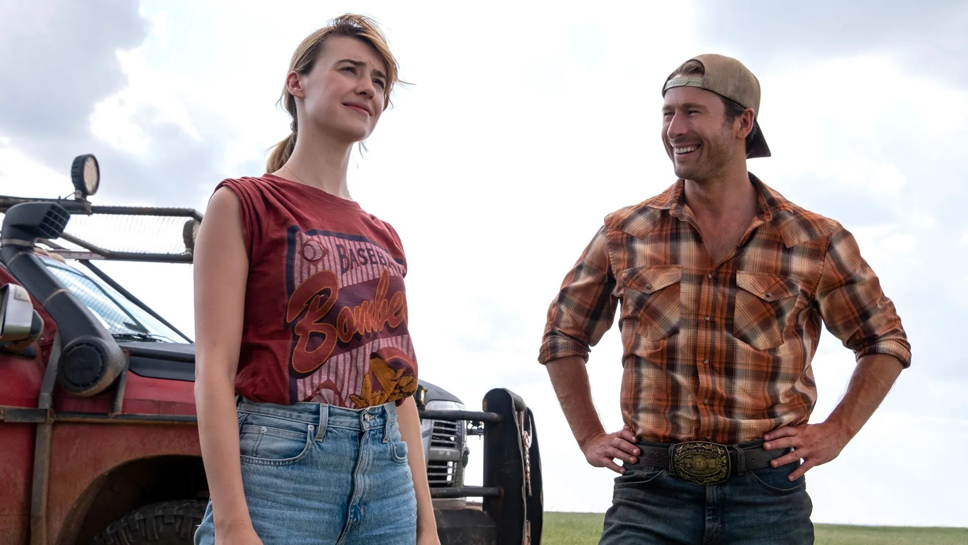 Una scena del film "Twisters" mostra due personaggi all'aperto in un ambiente rurale. A sinistra, una donna con una maglietta grafica rossa e pantaloncini di jeans guarda pensierosa in lontananza. A destra, un uomo con camicia a quadri, jeans e cappello da cowboy sorride sicuro di sé con le mani sui fianchi. Dietro di loro, è parcheggiato un robusto fuoristrada, a suggerire che si stanno preparando o riprendendo da un'avventura a caccia di tempeste. Il cielo luminoso e il paesaggio aperto contribuiscono all'atmosfera avventurosa del film.