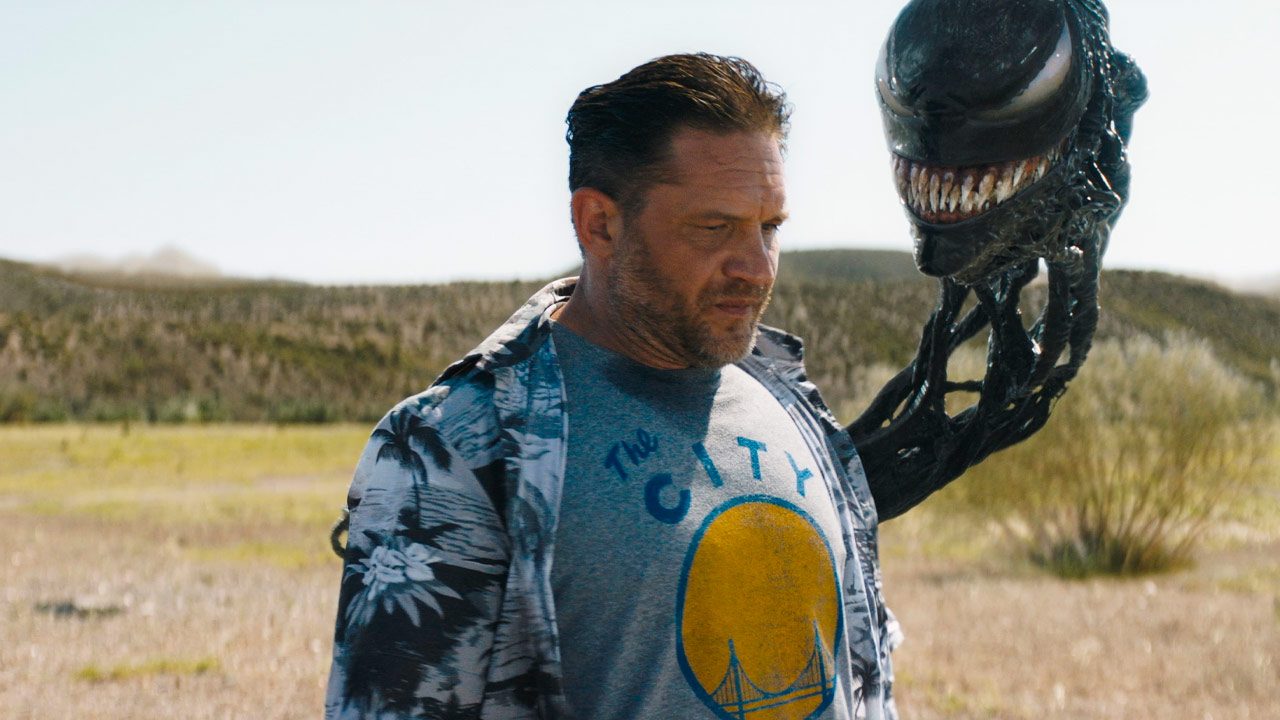 Una scena del film "Venom 3: The Last Dance" mostra Eddie Brock in piedi in un campo aperto e illuminato dal sole che indossa una camicia floreale casual sopra una maglietta con la scritta “The City” e un disegno a tema basket. Accanto a lui, il simbionte alieno Venom emerge dalla sua spalla, mostrando il suo ghigno minaccioso e pieno di denti. Le espressioni contrastanti - il contegno serio di Eddie e il ghigno sinistro di Venom - evidenziano la loro dinamica unica. Il paesaggio naturale sullo sfondo suggerisce un momento di riflessione o un imminente scontro.