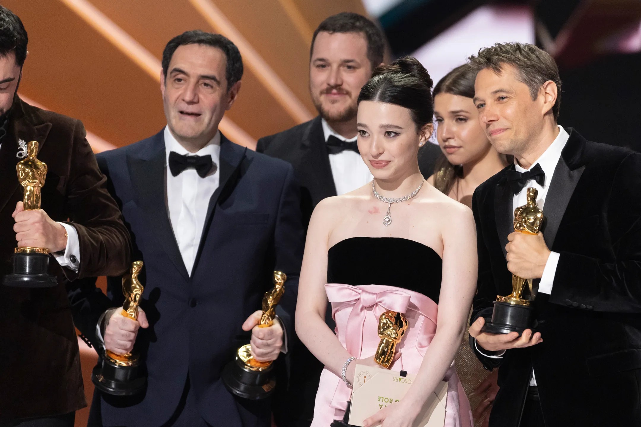 Il cast e la troupe di "Anora" sul palco degli Oscar 2025, visibilmente emozionati mentre stringono le loro statuette dorate. L'attrice Mikey Madison con un elegante abito nero e rosa tiene in mano l'Oscar e un foglio con la scritta 'Leading Role'. Accanto a lei, altri vincitori in smoking e abiti da sera sorridono e celebrano il traguardo raggiunto.