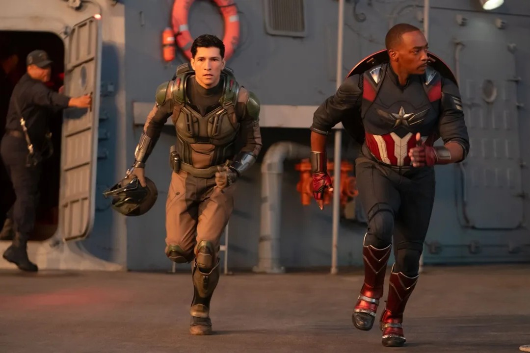 Scena d'azione tratta da 'Captain America: Brave New World'. Il nuovo Captain America, Sam Wilson, indossa il suo iconico costume con lo scudo sulle spalle mentre corre con espressione determinata. Al suo fianco, Joaquin Torres, in tenuta militare, stringe un casco mentre lo segue. Sullo sfondo, una nave militare con dettagli metallici e un marinaio che esce da una porta.