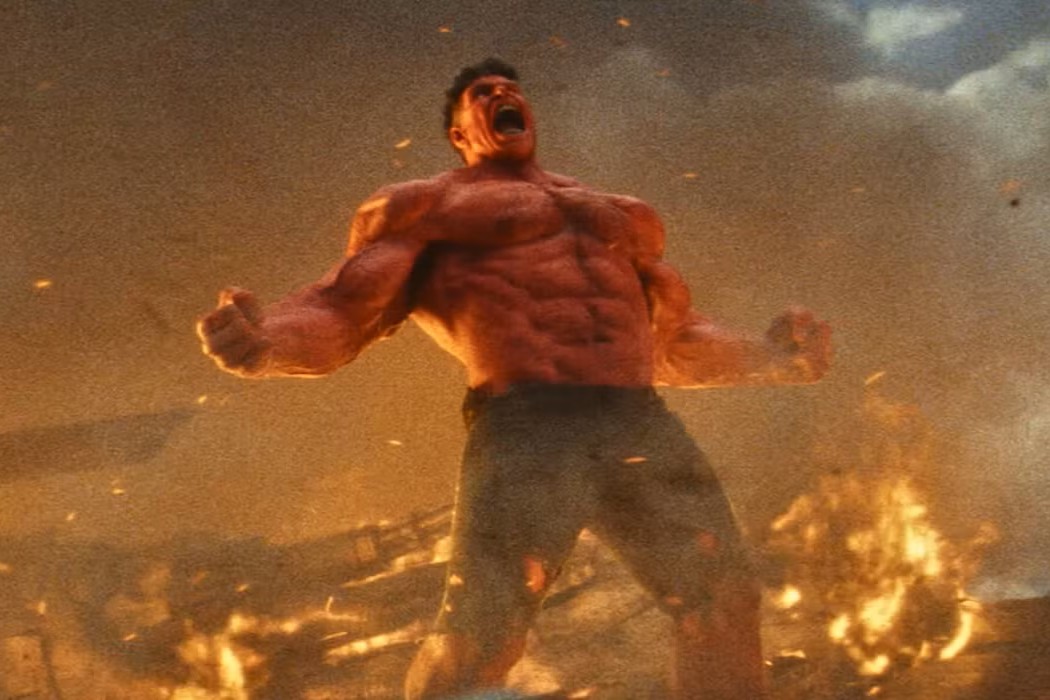 Scena tratta da "Captain America: Brave New World": Red Hulk, con il torso nudo e pantaloni scuri, urla con rabbia contro il cielo. Il personaggio, illuminato dalla luce dorata del fuoco, è circondato da fiamme e detriti. L'inquadratura drammatica cattura il momento di furia del soggetto, con la sua muscolatura in evidenza e i pugni chiusi, simboleggiando potenza e disperazione in un contesto di distruzione.