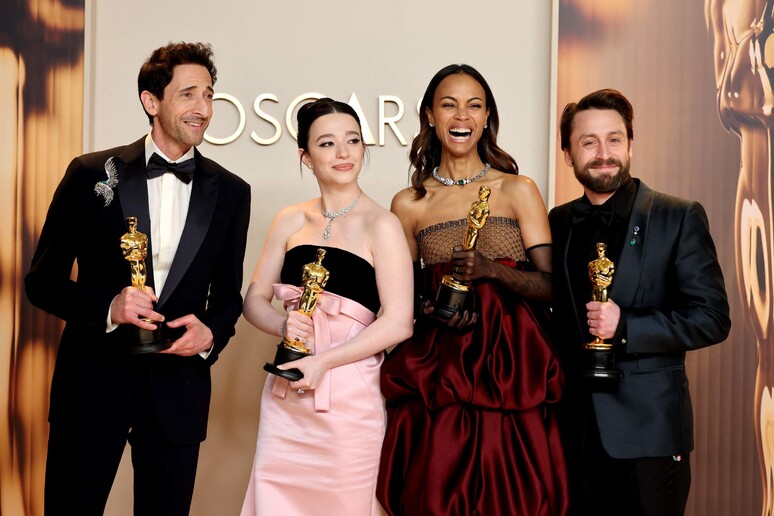 Quattro vincitori degli Oscar 2025 posano sorridenti nella sala stampa, tenendo in mano le loro statuette dorate. Indossano abiti eleganti: due uomini in smoking, una donna in un raffinato abito nero e rosa, e un'altra in un voluminoso vestito rosso scuro. Sullo sfondo, la scritta 'Oscars' e decorazioni dorate evocano l'atmosfera glamour della cerimonia.